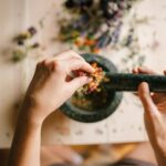 A close-up view of hands grinding herbs and flowers in a mortar and pestle on a wooden table.