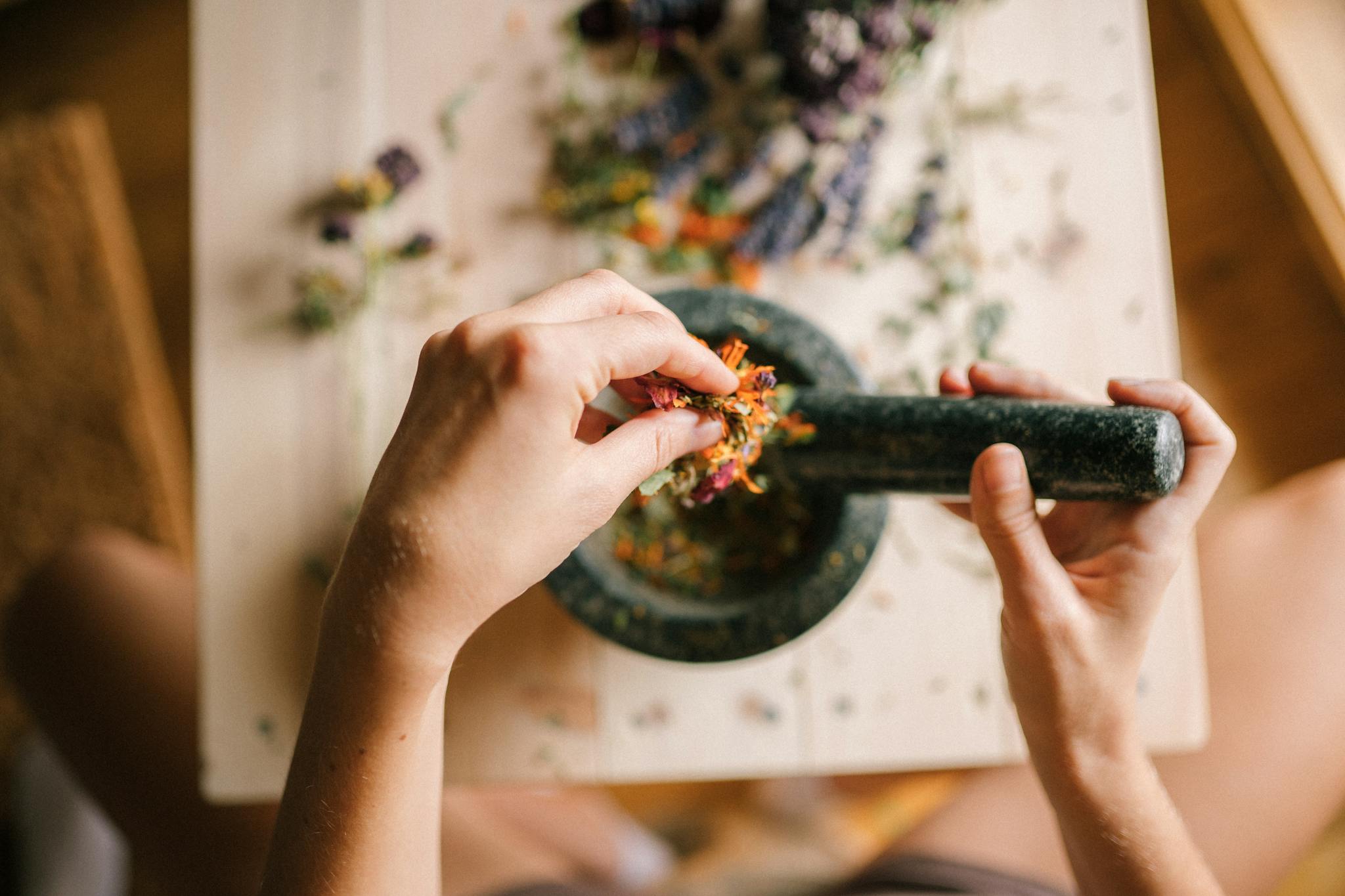 A close-up view of hands grinding herbs and flowers in a mortar and pestle on a wooden table.